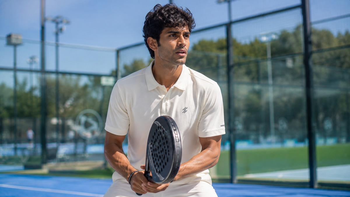 A man on a blue padel court wearing a cream polo t shirt with Dcyphr Shorts which looks an athlete on the ground. Fashion with Sports look. 