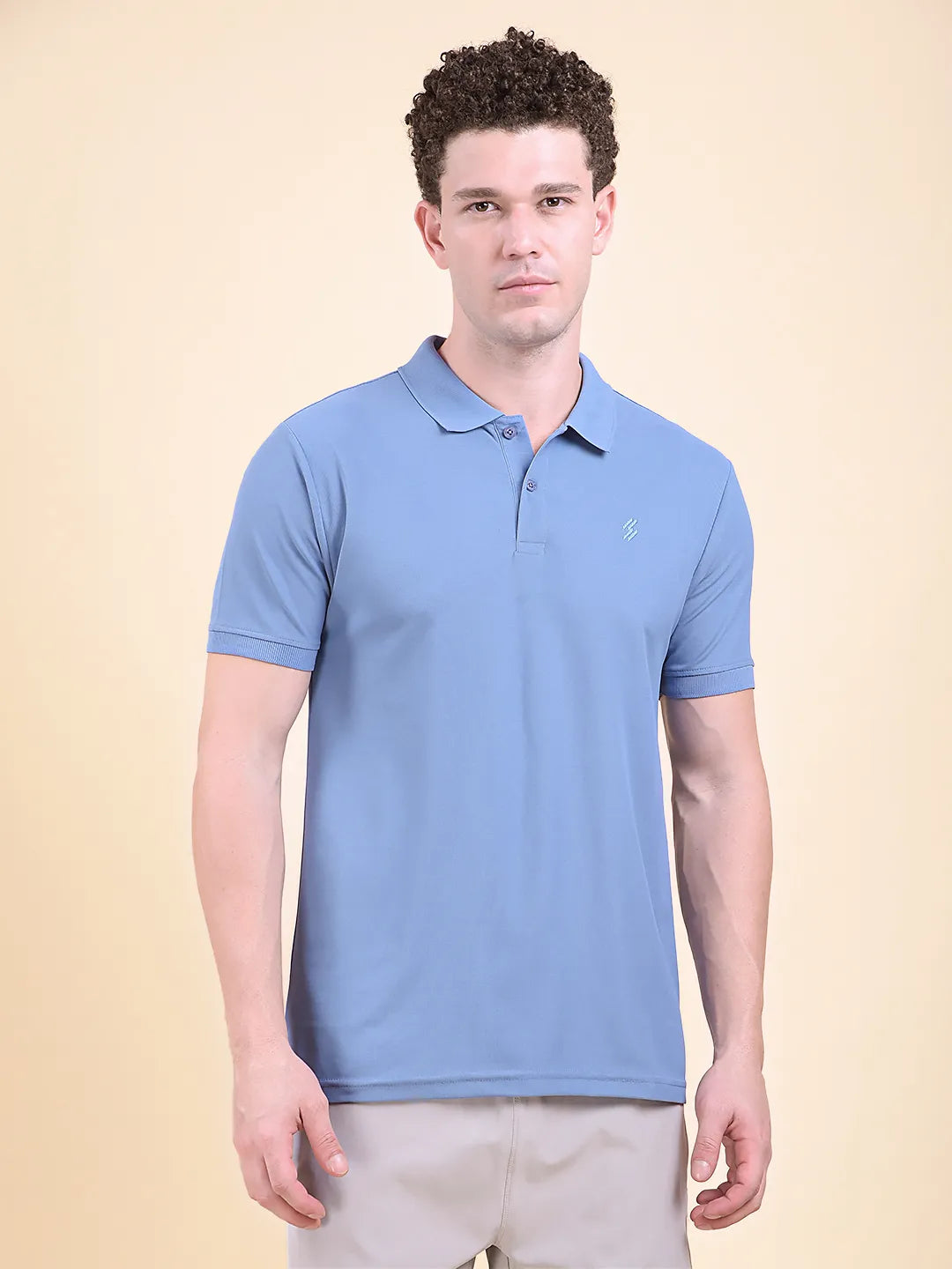 Man wearing a blue polo t-shirt  (DCYPHR Men's Element Polo - Denim Blue) against a beige background front facing towards camera.