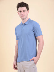 Man wearing a blue polo t-shirt  (DCYPHR Men's Element Polo - Denim Blue) against a beige background facing towards camera posing side ways.