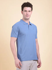Man wearing a blue polo t-shirt  (DCYPHR Men's Element Polo - Denim Blue) against a beige background front facing.