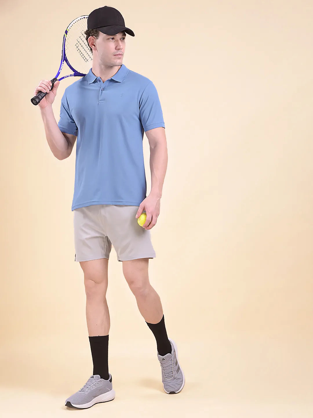 Man wearing a blue polo t-shirt  (DCYPHR Men's Element Polo - Denim Blue) holding a tennis racket and a ball against a beige background
