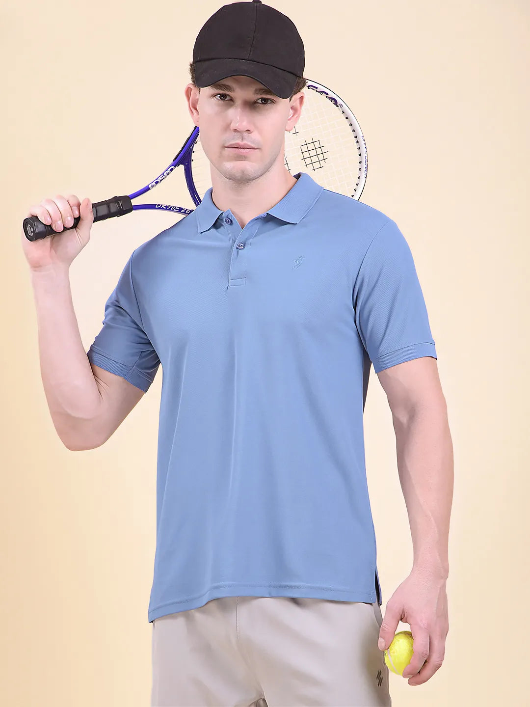 Man holding a tennis racket and ball, wearing a light blue polo t-shirt (DCYPHR Men's Element Polo - Denim Blue) and black cap on a beige background.
